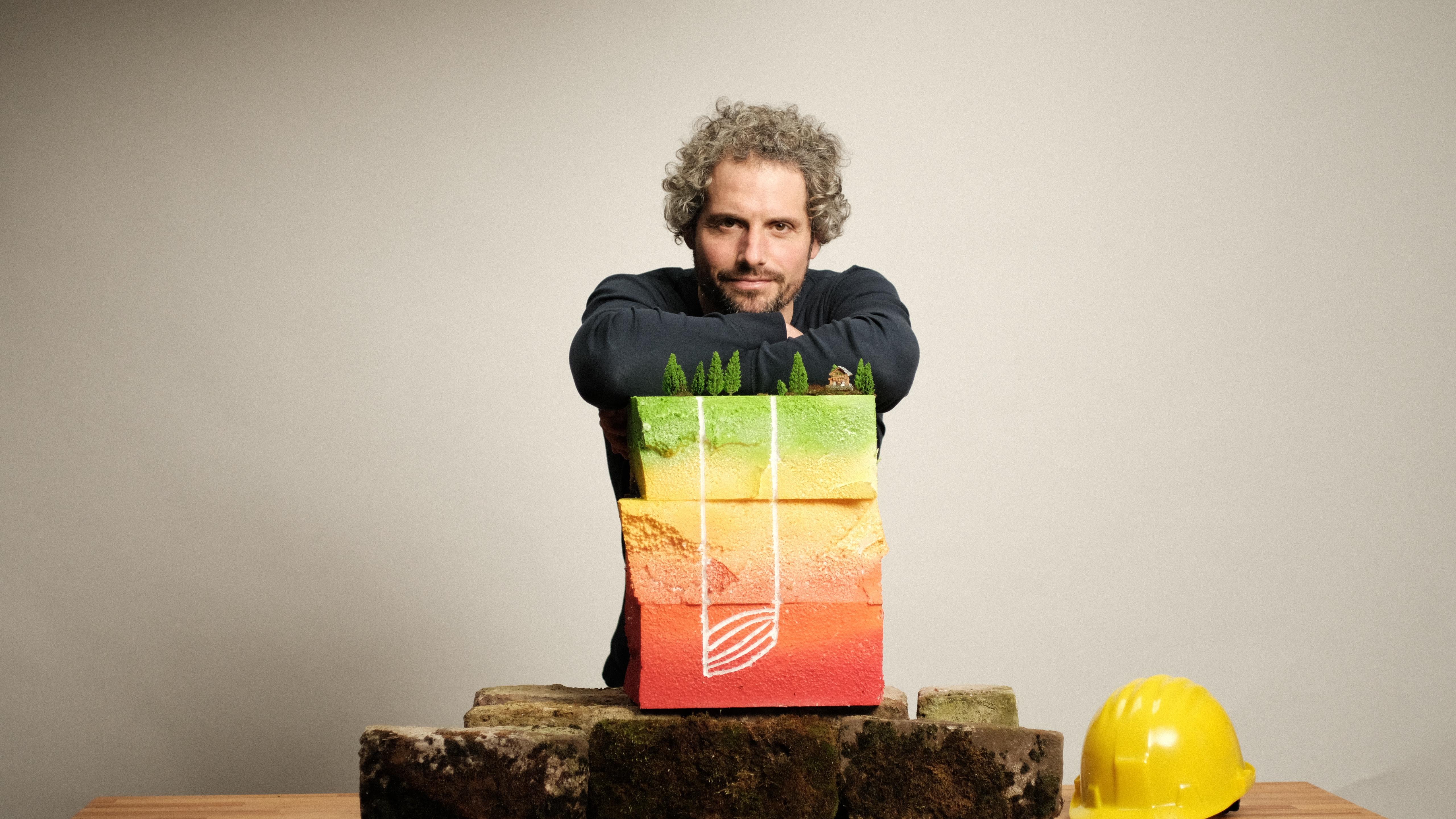 Ein Mann mit lockigem Haar - Tim Schauenberg, Klima- und Wirtschaftsjournalist und Moderator aus Köln - posiert mit einem farbigen Modell, das eine Landschaft darstellt, und hält es vor einem neutralen Hintergrund. Neben ihm liegt ein gelber Helm.
Es geht um neue Energien, grüne Energie , Nachhaltigkeit, Klima, Kreislaufwirtschaft und Tim Schauenberg, Journalist und Moderator aus Köln und Deutschland.