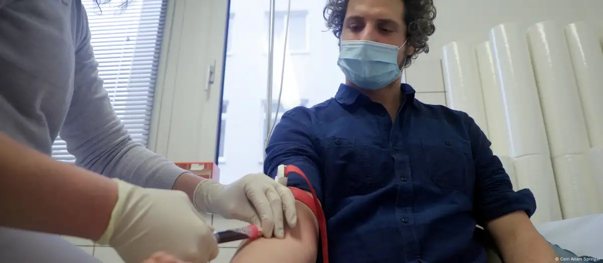 Tim Schauenberg, internationaler Moderator und Journalist aus Köln sitzt beim Arzt auf der Bank. Er streckt dem Arm aus während ihm eine Nadel in die Adern gestochen werden. Er trägt eine Maske. Die Probe dient dem Nachweis von Ewigkeitschemikalien in seinem Blut. Das ist Teil eines Recherhcehprojekts an dem er gearbeitet hat. 