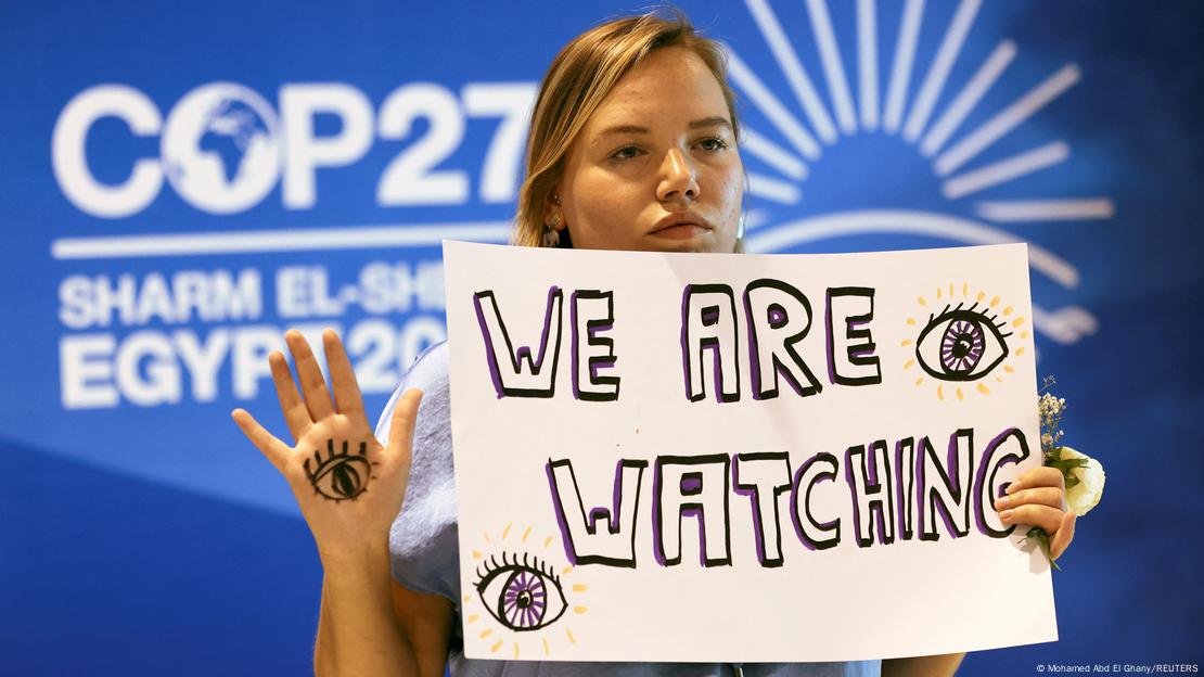 Eine Aktivistin steht bei der Klimakonferenz COP27 in Sharm Al Sheikh vor einem Plakat mit der Aufschrift COP27. Sie hält ein Schild hoch, darauf steht "We are watching". Auf ihrer hochgehaltenen Hand ist ein Auge gemalt.