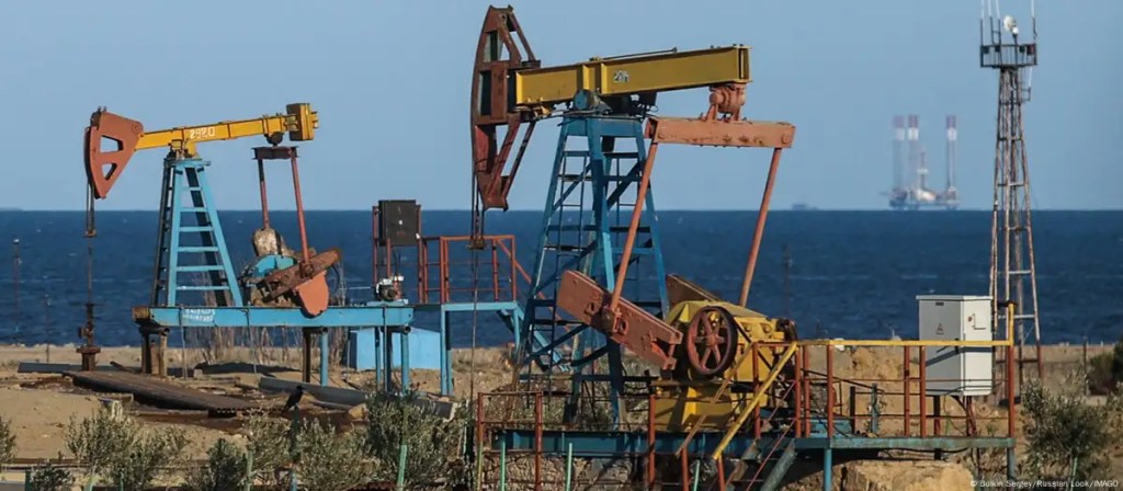 Auf einer trockene Ebene stehen mehrere Ölpumpen und pumpen Öl aus der Tiefe. Im Hintergrund das Meer, darauf ist eine Ölbohrinsel zusehen.