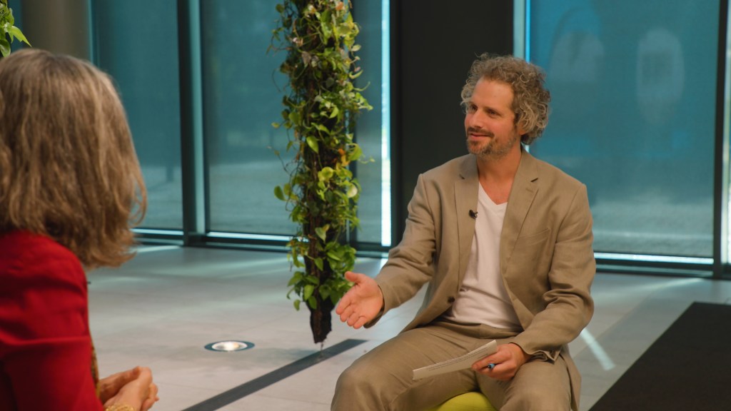 Journalist und Moderator im beigen Anzug führt ein professionelles Interview mit einer Gesprächspartnerin im roten Blazer.