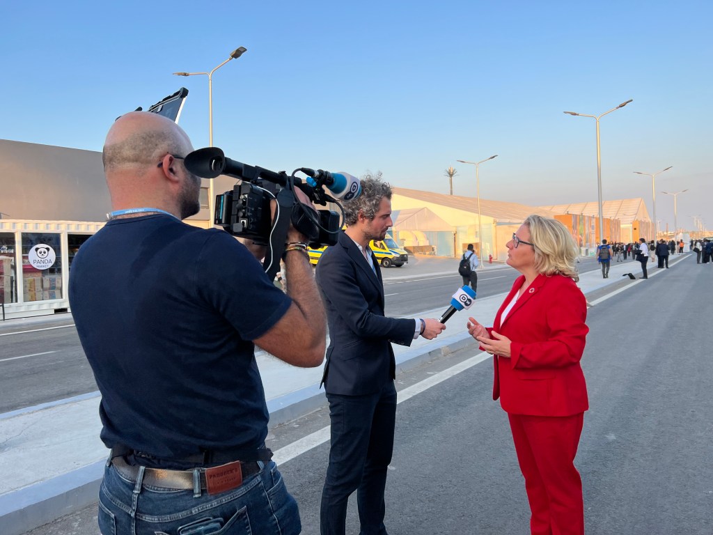 Tim Schauenberg, Experte und Klima- und Wirtschaftsjournalist, Moderator aus Köln, steht vor der Kamera auf der Klimakonferenz, bei der die Politik und Politiker mit Hilfe internationaler Diplomatie und Politik über Klimaneutralität und Emissionen verhandeln. Tim Schauenberg, der Journalist aus Köln, interviewt in diesem Moment Entwicklungsministerin Svenja Schulze, die mit einem roten Blaiser dem Moderator aus Köln Rede und Antwort steht in einer Live-Schalte und Moderation aus in Ägypten. In der Hand hält Tim Schauenberg ein Mikrofon , darauf zu sehen das Deutsche Welle Logo im Hintergrund die Abendsonne von Sharm Al Sheik der Tourismusort in Ägpyten. Tim Schauenberg ist Experte in der Berichterstattung zu Themen der Kreislaufwirtschaft und anderen Nachhaltigkeitsthemen, rund um Wirtschaft und Klima.