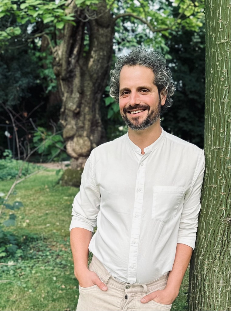 Tim Schauenberg, Klima- und Wirtschaftsjournalist und Moderator aus Köln, lehnt an einem Baum, er trägt ein weißes Hemd und lächelt in die Kamera. Im Hintergrund stehen grüne Bäume und grüne Elemente, die das Bild um Tim Schauenberg, Journalist aus Köln und Moderator für Klimathemen in ein Licht der Nachhaltigkeit rücken und grün einbetten