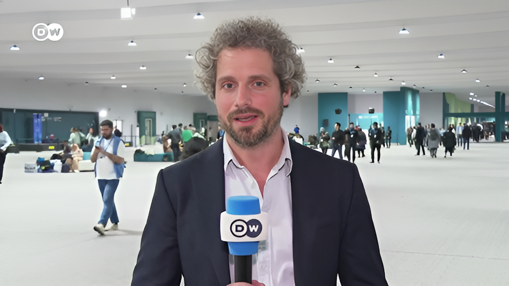 Der Journalist für Klima, Wirtschaft und Nachhaltigkeit, Tim Schauenberg steht mit einem Mikrofon vor der Kamera und spricht für das Fernsehen als Moderator in die Kamera. Hinter ihm eine Halle in der mehrere Menschen zu Umweltthemen beraten.